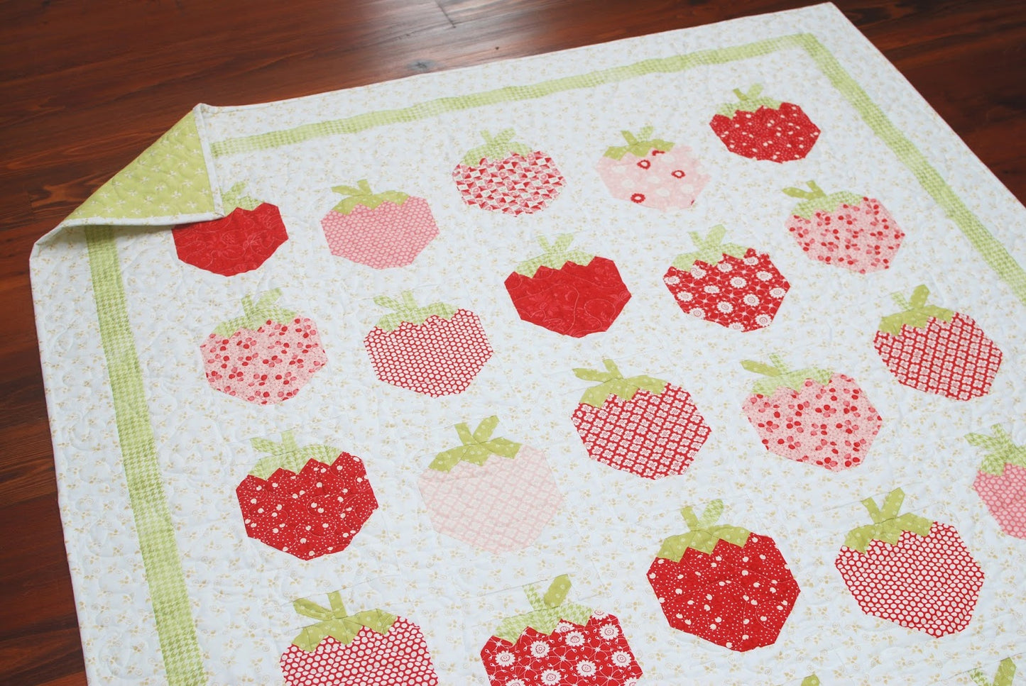 A quilted textile featuring a pattern of red and pink strawberries on a white background with a light green border, shown laid out on a dark wood floor with one corner folded back.
Photo by ThePatternBasket.com [additional_image_link]