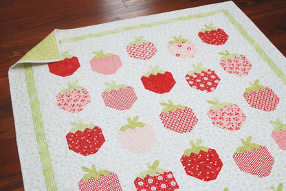 A quilted textile featuring a pattern of red and pink strawberries on a white background with a light green border, shown laid out on a dark wood floor with one corner folded back.
Photo by ThePatternBasket.com [additional_image_link]