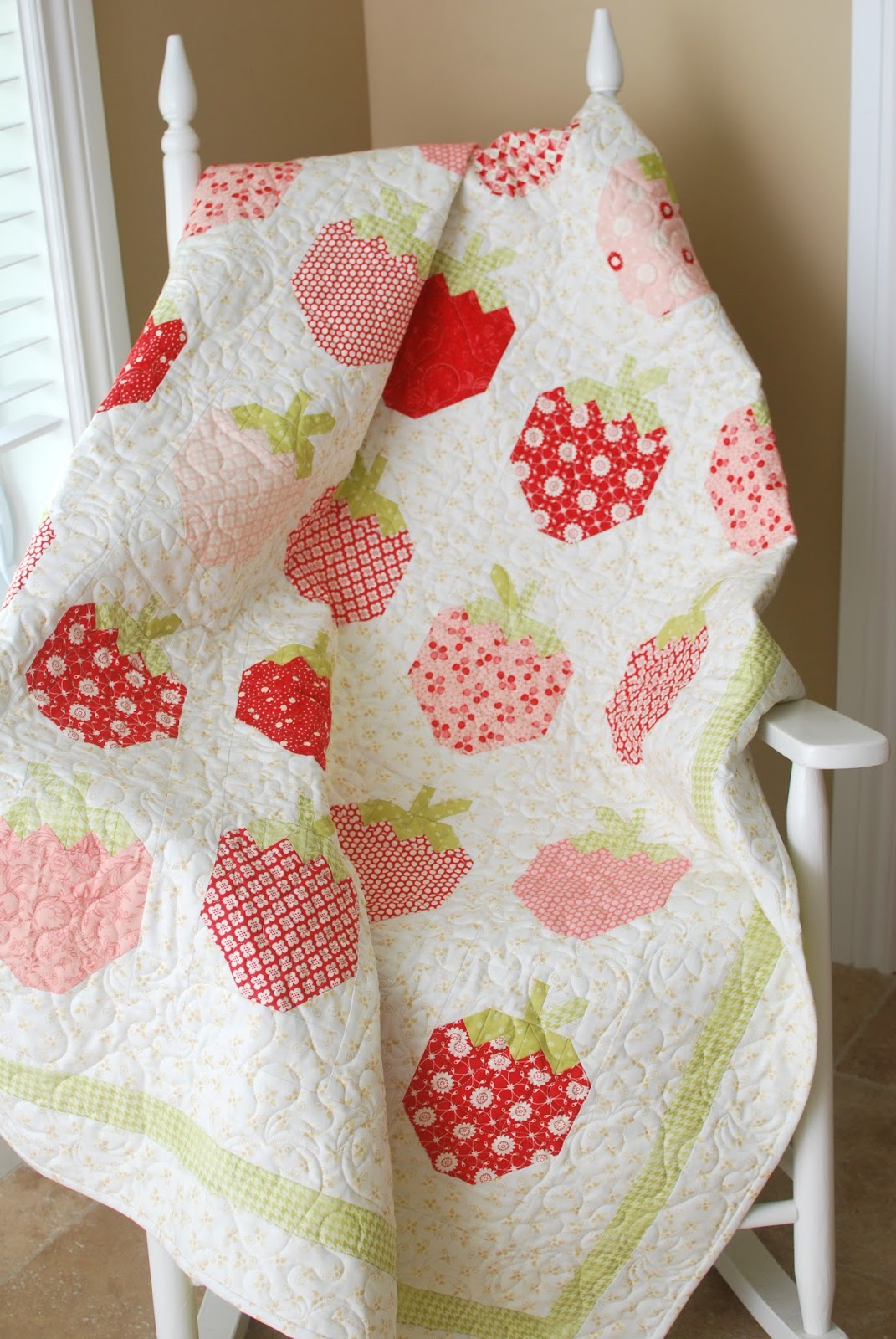 A quilted baby blanket with a strawberry pattern in shades of red, pink, and green, draped over a white wooden chair in a home setting.
Photo by ThePatternBasket.com [additional_image_link]