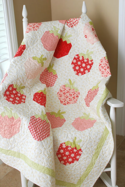 A quilted baby blanket with a strawberry pattern in shades of red, pink, and green, draped over a white wooden chair in a home setting.
Photo by ThePatternBasket.com [additional_image_link]