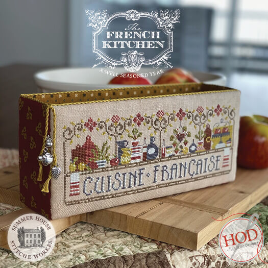 A decorative rectangular cross-stitch sampler box with a French kitchen theme, displayed on a wooden cutting board with apples in a kitchen setting. The sampler features embroidered motifs and the text 'CUISINE FRANÇAISE'.
Photo by HandsOnDesign.biz [image_link]