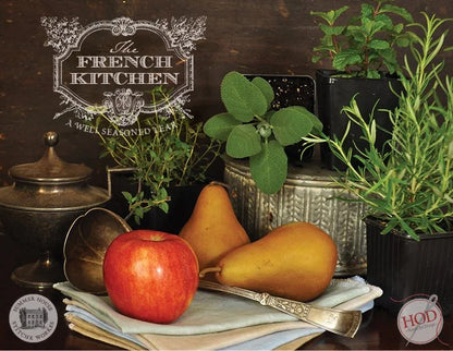 A styled still life scene with a red apple, two pears, green herbs, and potted plants arranged on a dark wooden table with a metal bowl, folded cloth, and silver spoon. A decorative sign reads 'The French Kitchen' and there are circular 'HOD' and 'Summer House Stitche Workes' logos in the corners.
Photo by HandsOnDesign.biz [additional_image_link]