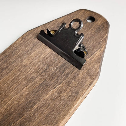A close-up of a dark brown wooden board with a black metal binder clip attached near the top, set against a plain white background.
Photo by HandsOnDesign.biz [additional_image_link]