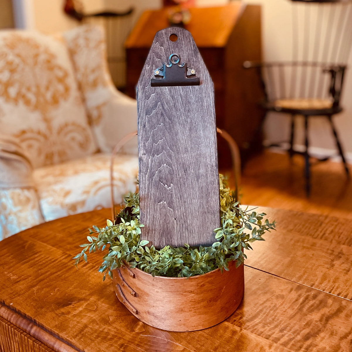 A rustic wooden sign holder with a weathered gray wood plaque and a small metal clip, displayed in a copper-colored wooden basket filled with green foliage on a wooden table, set in a cozy living room environment.
Photo by HandsOnDesign.biz [additional_image_link]
