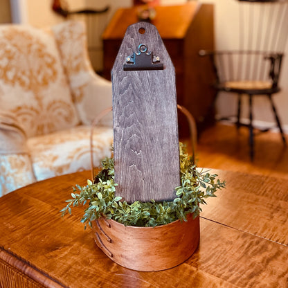 A rustic wooden sign holder with a weathered gray wood plaque and a small metal clip, displayed in a copper-colored wooden basket filled with green foliage on a wooden table, set in a cozy living room environment.
Photo by HandsOnDesign.biz [additional_image_link]