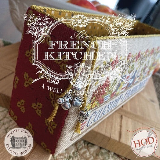 A close-up of a decorative fabric-covered box or case with a red side panel, multicolor floral patterned front, gold trim, and tassel zipper pull. Large overlaid text reads 'The French Kitchen' with smaller text 'A Well Seasoned Year'.
Photo by HandsOnDesign.biz [additional_image_link]