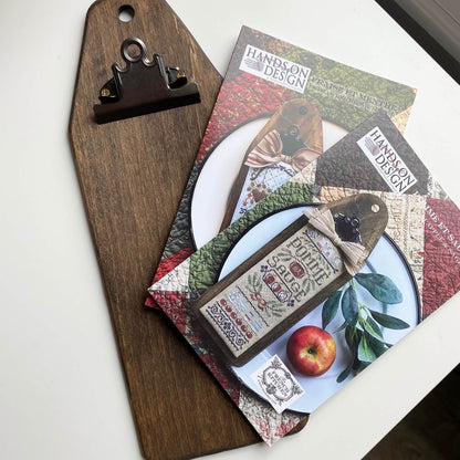 A styled flat-lay of a wooden cutting board with a dark metal clip, featuring printed craft paper sheets and tags with the brand name 'Hands on Design' and rustic autumn-themed illustrations.
Photo by HandsOnDesign.biz [additional_image_link]