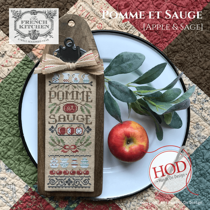 A decorative wooden cross-stitch sampler tag with a burlap bow and metal clasp, featuring the words “Pomme et Sauge” and apple-and-sage motifs. The tag is styled on a white plate with a red apple and green leaves, over a patchwork quilt background.
Photo by HandsOnDesign.biz [additional_image_link]