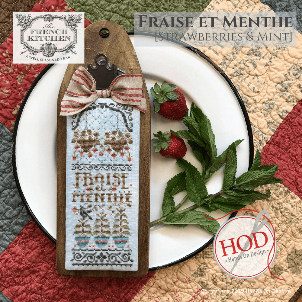 A decorative wooden cross-stitch sampler tag with a stitched French design reading 'Fraise et Menthe' is displayed on a white plate with strawberries and mint leaves. The tag has a striped fabric bow and a metal clasp.
Photo by HandsOnDesign.biz [additional_image_link]
