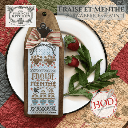 A decorative wooden cross-stitch sampler tag with a stitched French design reading 'Fraise et Menthe' is displayed on a white plate with strawberries and mint leaves. The tag has a striped fabric bow and a metal clasp.
Photo by HandsOnDesign.biz [additional_image_link]