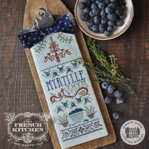 A finished cross-stitch sampler titled 'MYRTILLE THYM' on a wooden board, styled with a blue polka-dot bow, a clipboard clip, a bowl of blueberries, and fresh herbs on a dark wood background. The 'The French Kitchen' and 'Summer House Stitche Workes' logos are visible in the corners.
Photo by HandsOnDesign.biz [additional_image_link]