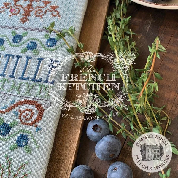 A close-up styled flat-lay of a cross-stitch sampler on a wooden board, with blueberries and fresh thyme sprigs. A large semi-transparent 'The French Kitchen' logo is overlaid across the center, and a circular 'Summer House Stitche Workes' seal appears in the lower right.
Photo by HandsOnDesign.biz [additional_image_link]