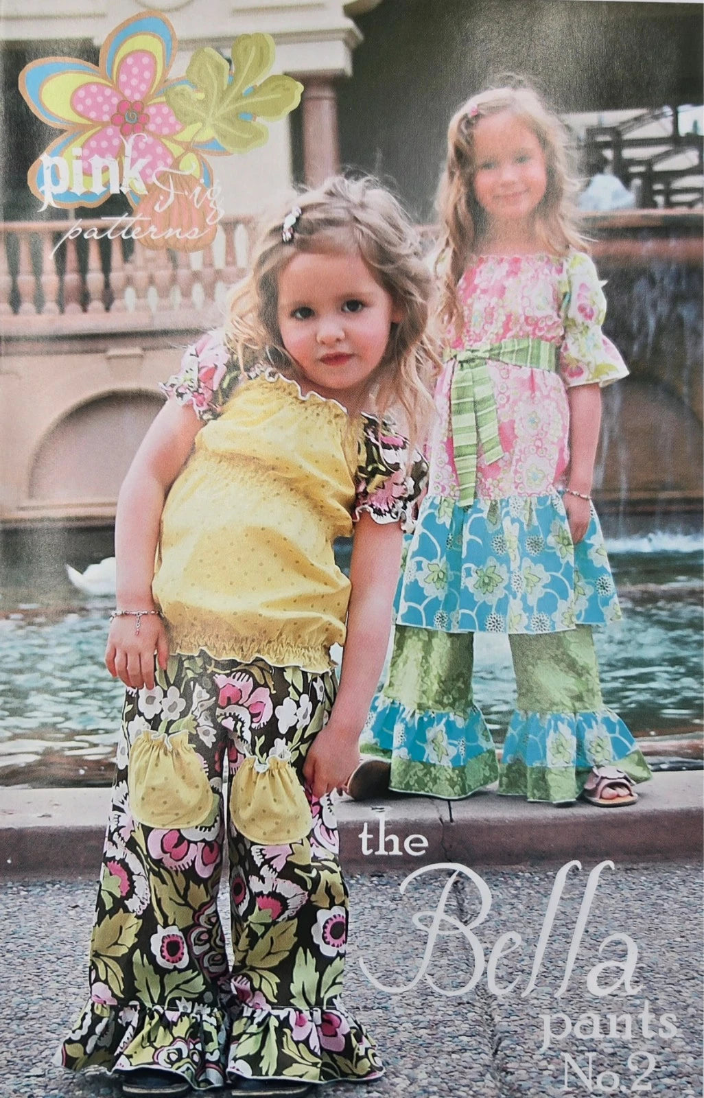 A vintage-style catalog or pattern cover showing two young girls outdoors in front of a fountain. The girl in front wears a yellow top with floral bell-bottom pants, and the girl behind wears a matching floral top and green patterned pants.
Photo by Pink Fig Design [image_link]