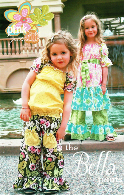 Two young girls modeling floral ruffled pants and matching tops in an outdoor setting with a fountain in the background. The image features a 'pink patterns' logo in the top left and 'the Bella pants No. 2
Photo by Pink Fig Patterns [additional_image_link]