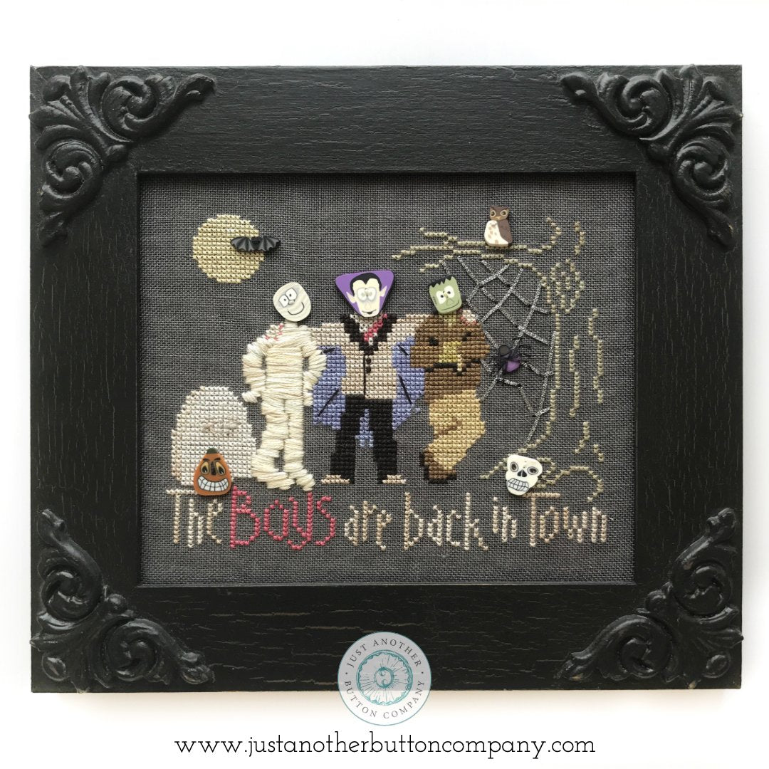 A framed cross-stitch artwork featuring three Halloween characters—a mummy, a vampire, and Frankenstein's monster—on a dark gray fabric background. The stitched text reads 'The Boys are back in town,' with small spider, moon, and skull details.
Photo by JustAnotherButtonCompany.com [additional_image_link]