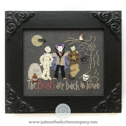 A framed cross-stitch artwork featuring three Halloween characters—a mummy, a vampire, and Frankenstein's monster—on a dark gray fabric background. The stitched text reads 'The Boys are back in town,' with small spider, moon, and skull details.
Photo by JustAnotherButtonCompany.com [additional_image_link]