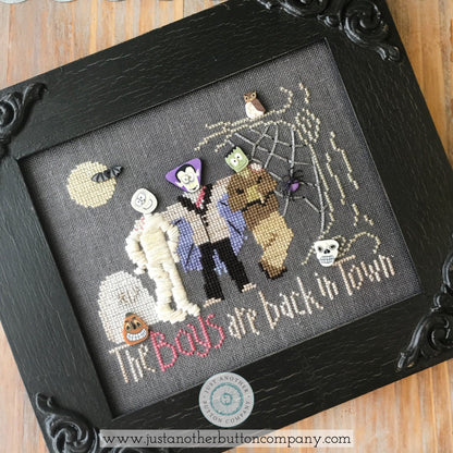 A framed cross-stitch artwork featuring three cartoon-style Halloween characters with spiderwebs, a moon, and the text 'The Boys are back in Town.' The piece is shown in a black ornate frame on a wooden surface.
Photo by JustAnotherButtonCompany.com [additional_image_link]