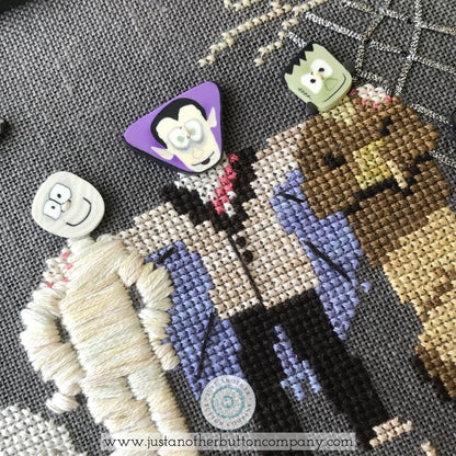 A close-up of a cross-stitch design featuring characters from The Addams Family, including Uncle Fester, Morticia, Wednesday Addams, and the Addams Bear, on dark gray Aida cloth. Several small decorative buttons are placed on top of the finished stitching.
Photo by JustAnotherButtonCompany.com [additional_image_link]