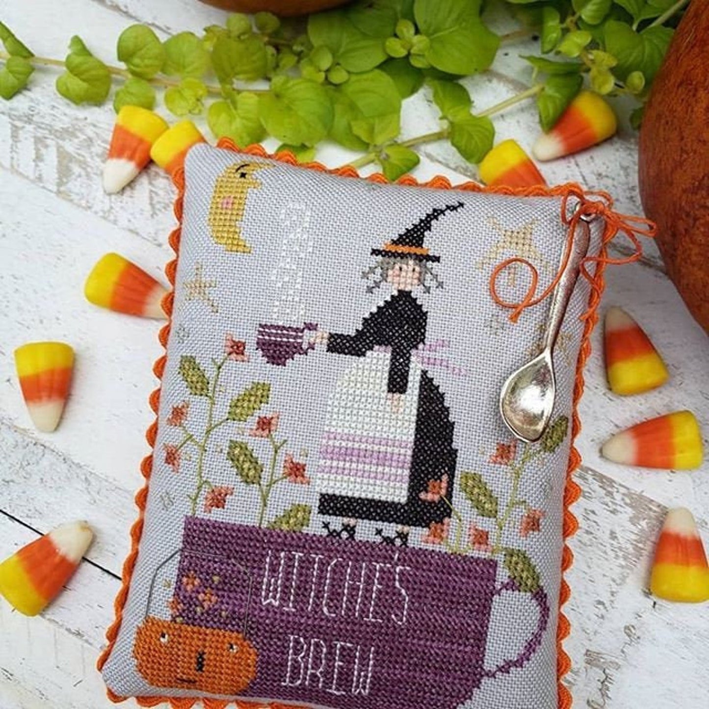 Cross-stitched pillow with witch design and 'Witch's Brew' text, surrounded by candy corn and leaves on a wooden surface.
[additional_image_link]