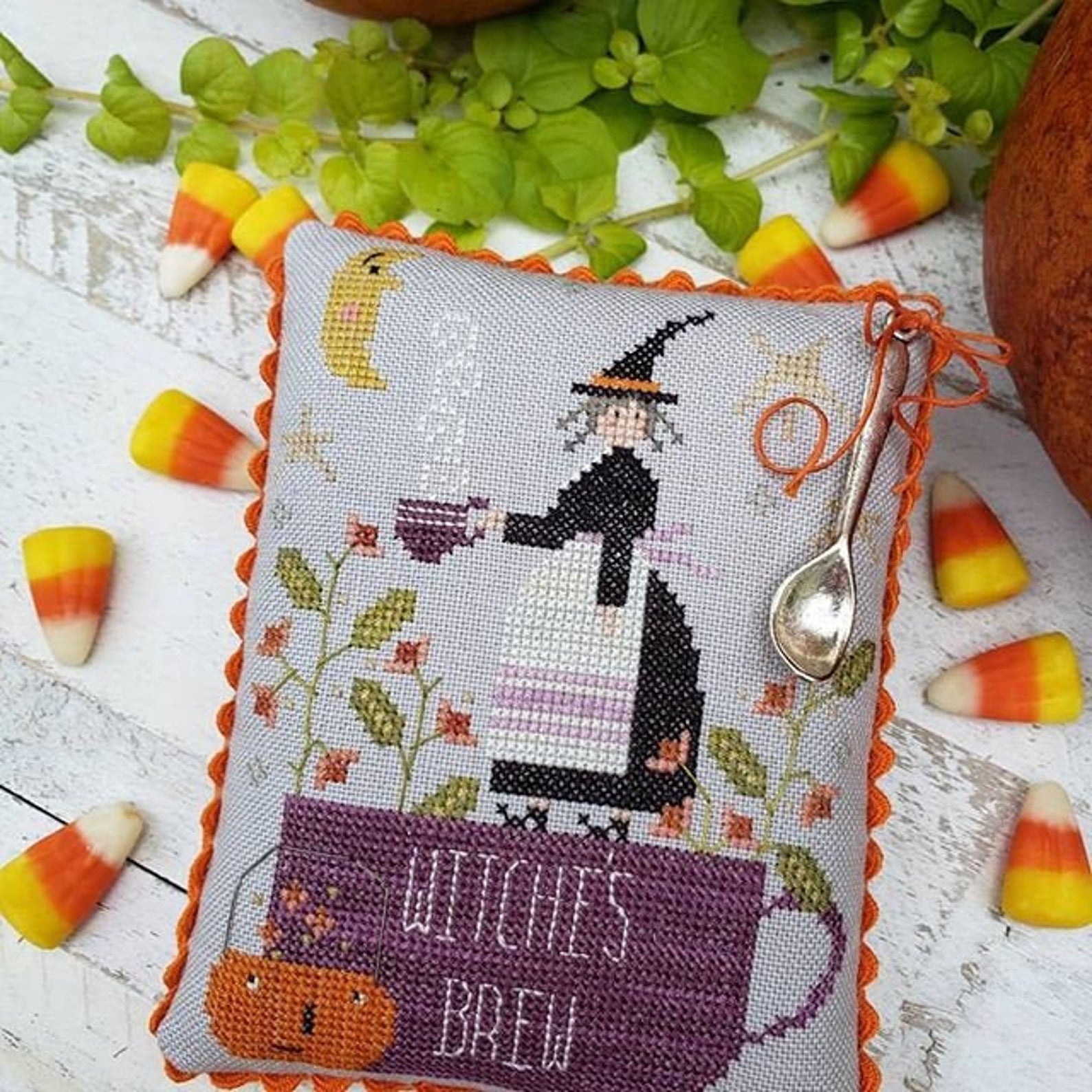 Cross-stitched pillow with witch design and 'Witch's Brew' text, surrounded by candy corn and leaves on a wooden surface.
[additional_image_link]