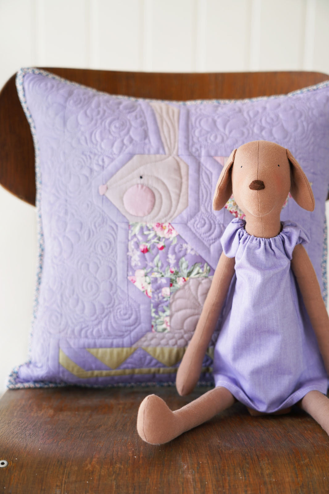 A lavender quilted throw pillow with a rabbit applique design sits on a wooden chair next to a plush stuffed dog doll wearing a matching lavender dress.
Photo by Tildafabrics.com [additional_image_link]