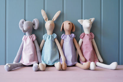 Four handmade fabric animal dolls sitting side by side against a blue paneled background. The dolls resemble a mouse, a rabbit, a dog, and a cat, each wearing a different pastel-colored dress in pink, light blue, lavender, and dusty pink.
Photo by Tildafabrics.com [additional_image_link]