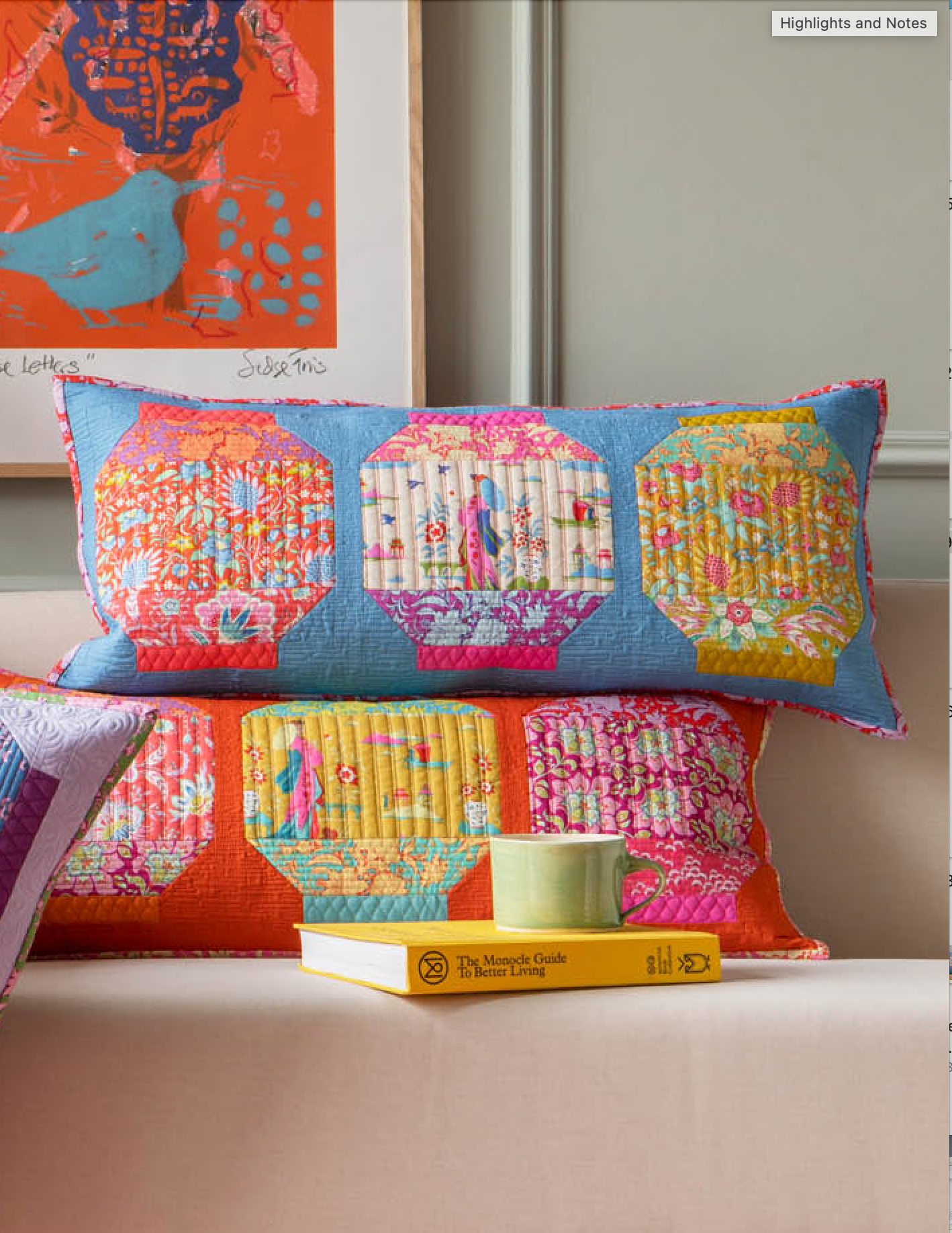 Two decorative lumbar pillows with colorful patchwork floral and geometric quilted panels in blue, orange, pink, yellow, and white, styled on a sofa with a book and mug in a home setting.