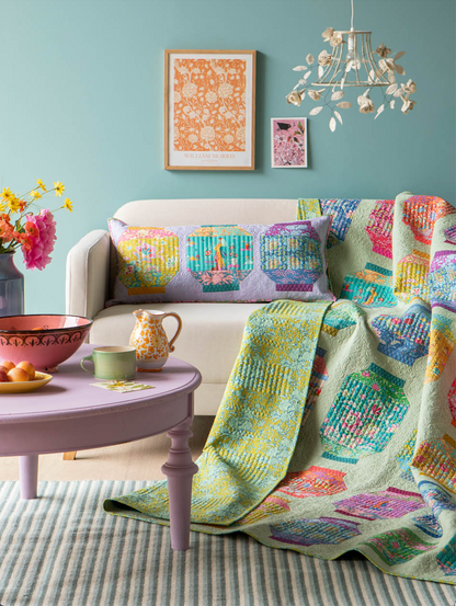 A cozy living room scene featuring a colorful quilted throw blanket draped over a cream sofa, a matching patterned lumbar pillow, and a lavender side table with flowers and a teapot.
Photo by Tildafabrics.com [additional_image_link]