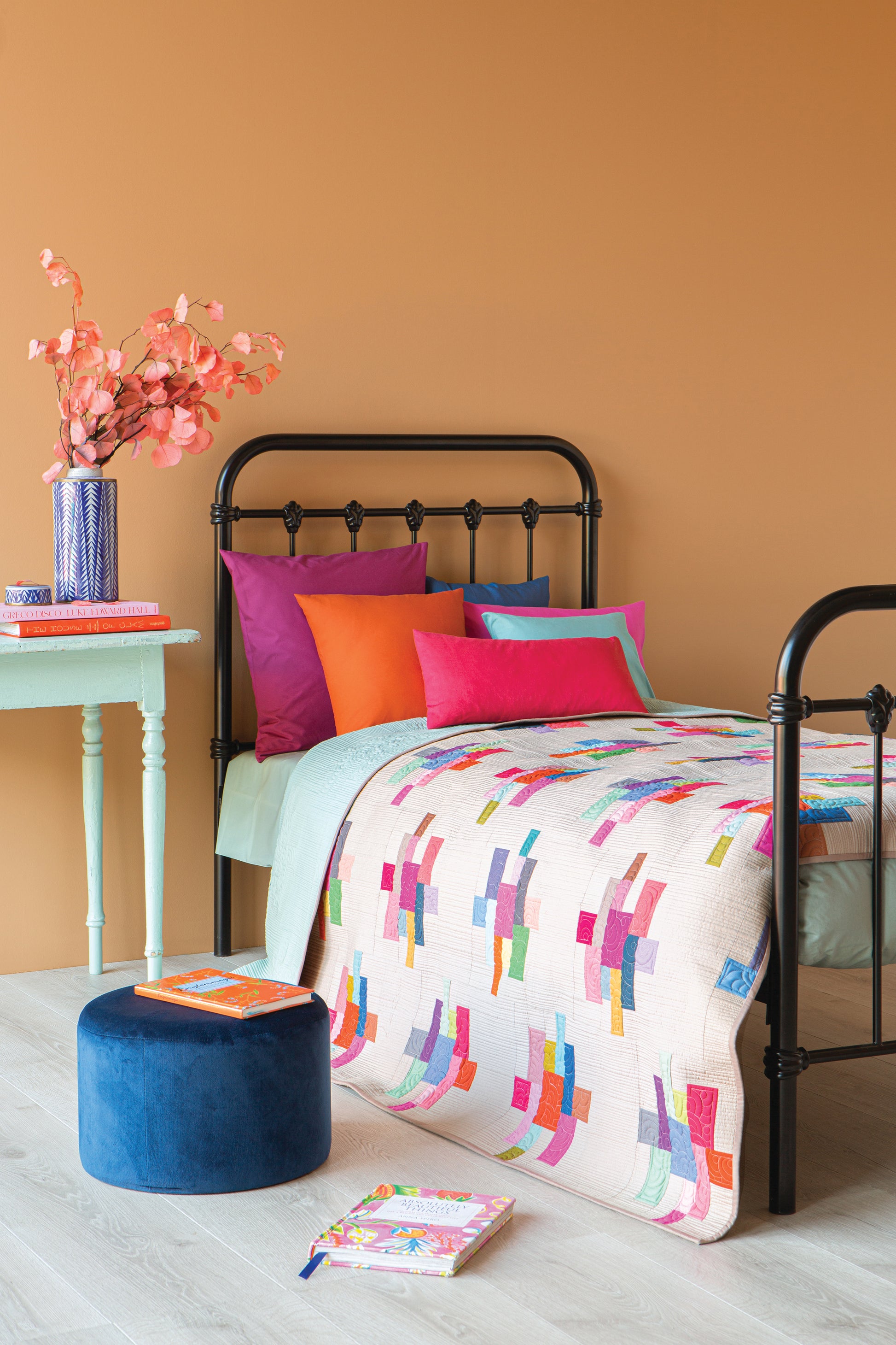 A styled single bed in a bedroom setting with a black metal frame, featuring a white duvet cover with a colorful geometric pattern, bright pink, purple, and orange pillows, a light green side table with flowers, and a blue velvet ottoman against a warm orange wall.
Photo by Tildafabrics.com [additional_image_link]