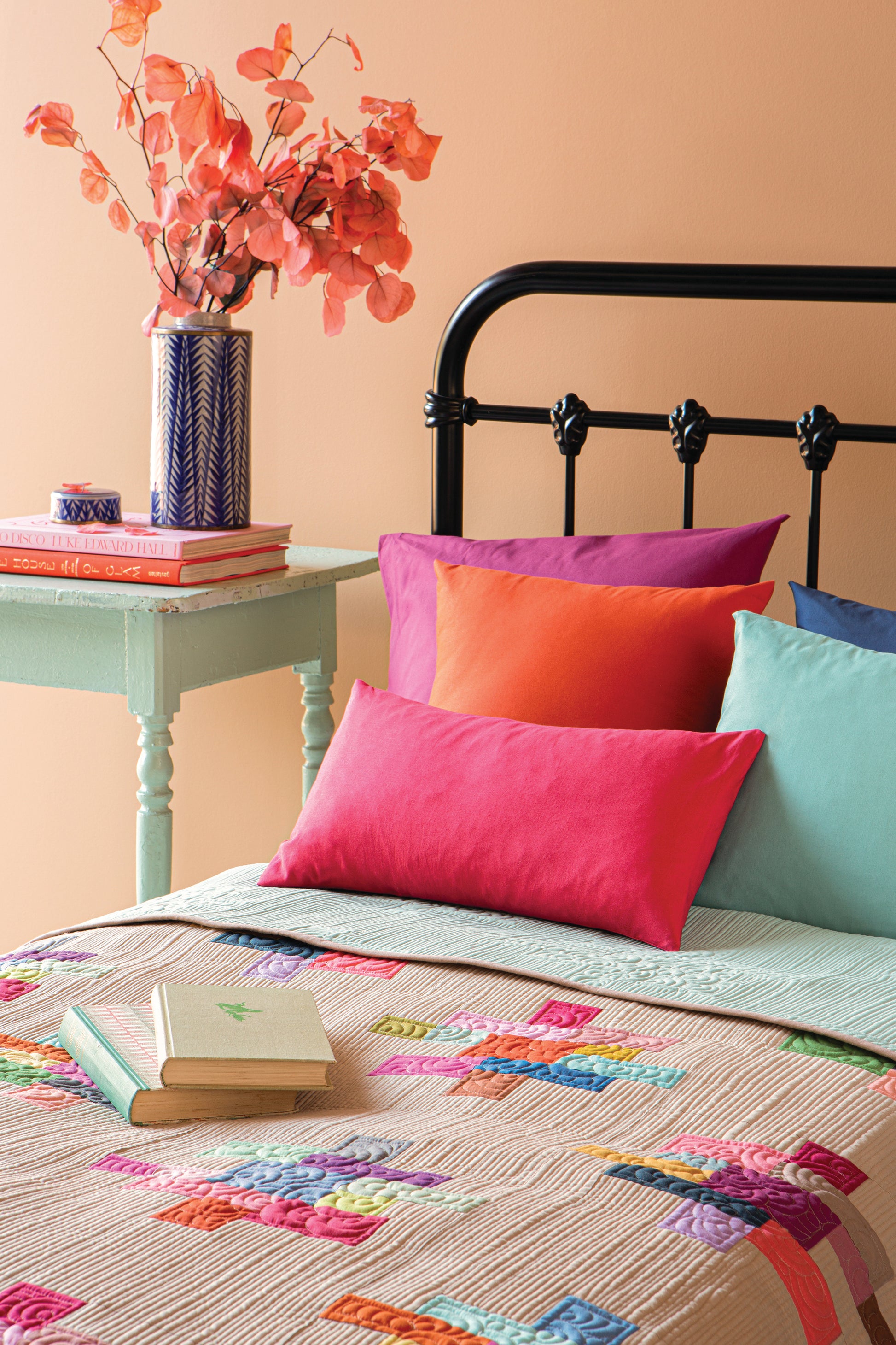 A styled bedroom scene with a bed made up with a multicolored patchwork quilt, bright pink, purple, red, and mint pillows, a black metal headboard, and a mint green bedside table holding a vase of red flowers and books.
Photo by Tildafabrics.com [additional_image_link]