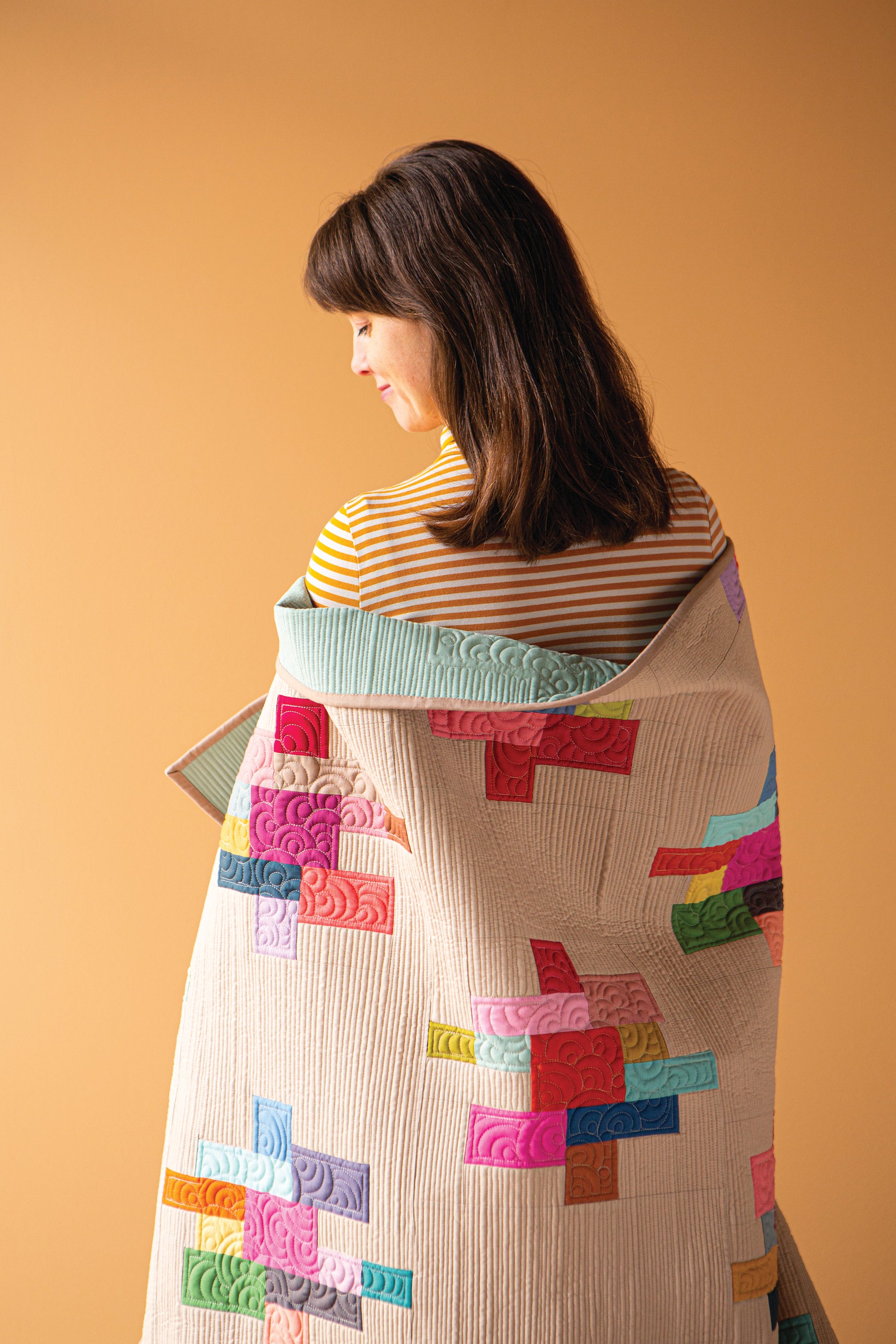 A woman is shown from behind wearing a quilted blanket or wrap with a colorful speckled patchwork design over a striped shirt, against a solid peach background. A logo in the top left reads 'Tilda SOLDS Colour Speckles Quilt'.
Photo by Tildafabrics.com [additional_image_link]