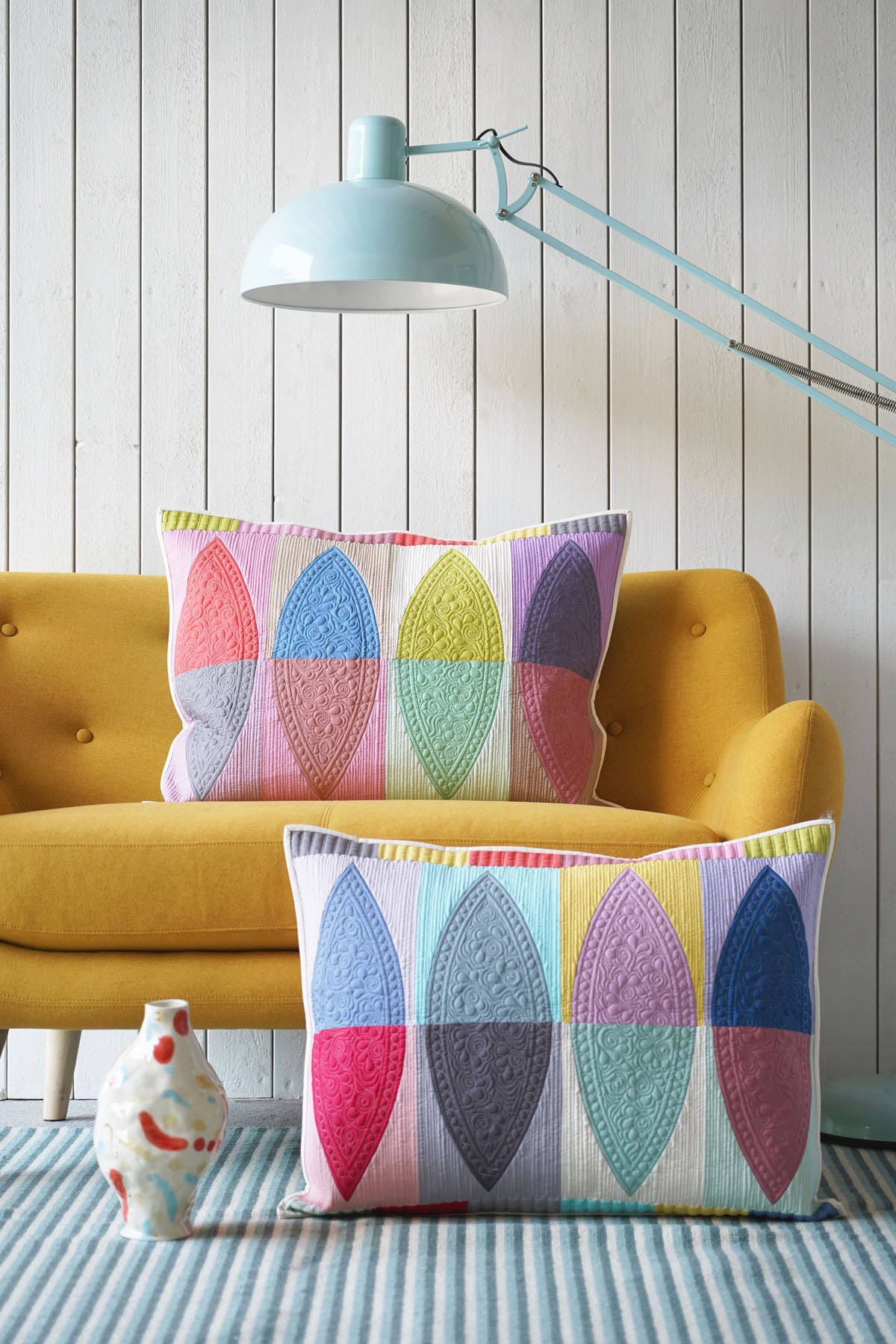Two decorative quilted pillows with a colorful geometric leaf pattern are styled in a living room setting, one resting on a mustard yellow armchair and the other on a blue-and-white striped rug in front of it. A light blue floor lamp and a white paneled wall are in the background.
Photo by Tildafabrics.com [image_link]