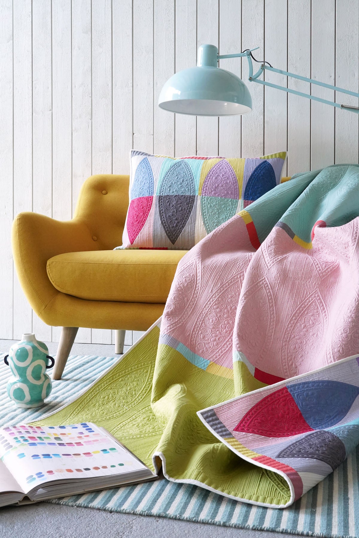 A styled interior scene showing a pastel quilt draped over a chair and floor, with a matching patterned throw pillow on a yellow armchair, an open book, a small vase, and a light blue wall lamp against white paneled walls.
Photo by Tildafabrics.com [additional_image_link]