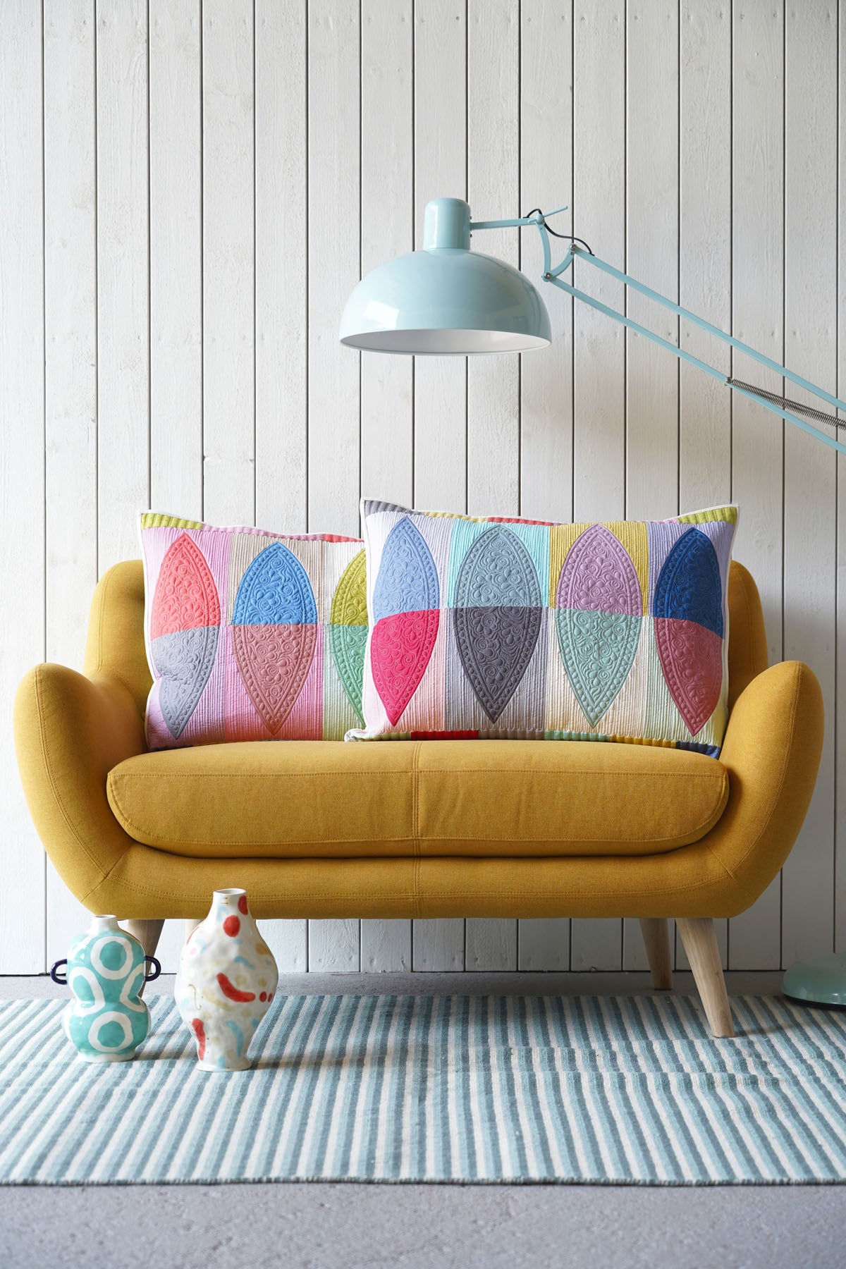 A yellow upholstered loveseat with two colorful quilted pillows, set against a white wood-paneled wall with a light blue floor lamp and striped rug.
Photo by Tildafabrics.com [additional_image_link]