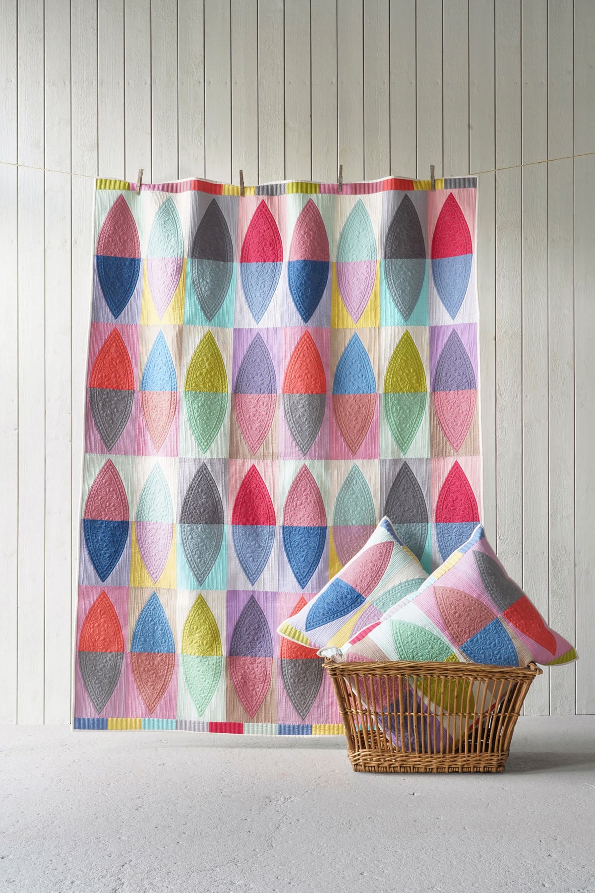 A colorful photo featuring a large quilt and two matching pillows, both with a repeating geometric leaf-like pattern in pink, blue, yellow, green, and gray on a white base. The hanging is clipped to a string against a white paneled wall, with a wicker basket holding two pillows in the foreground.
Photo by Tildafabrics.com [additional_image_link]
