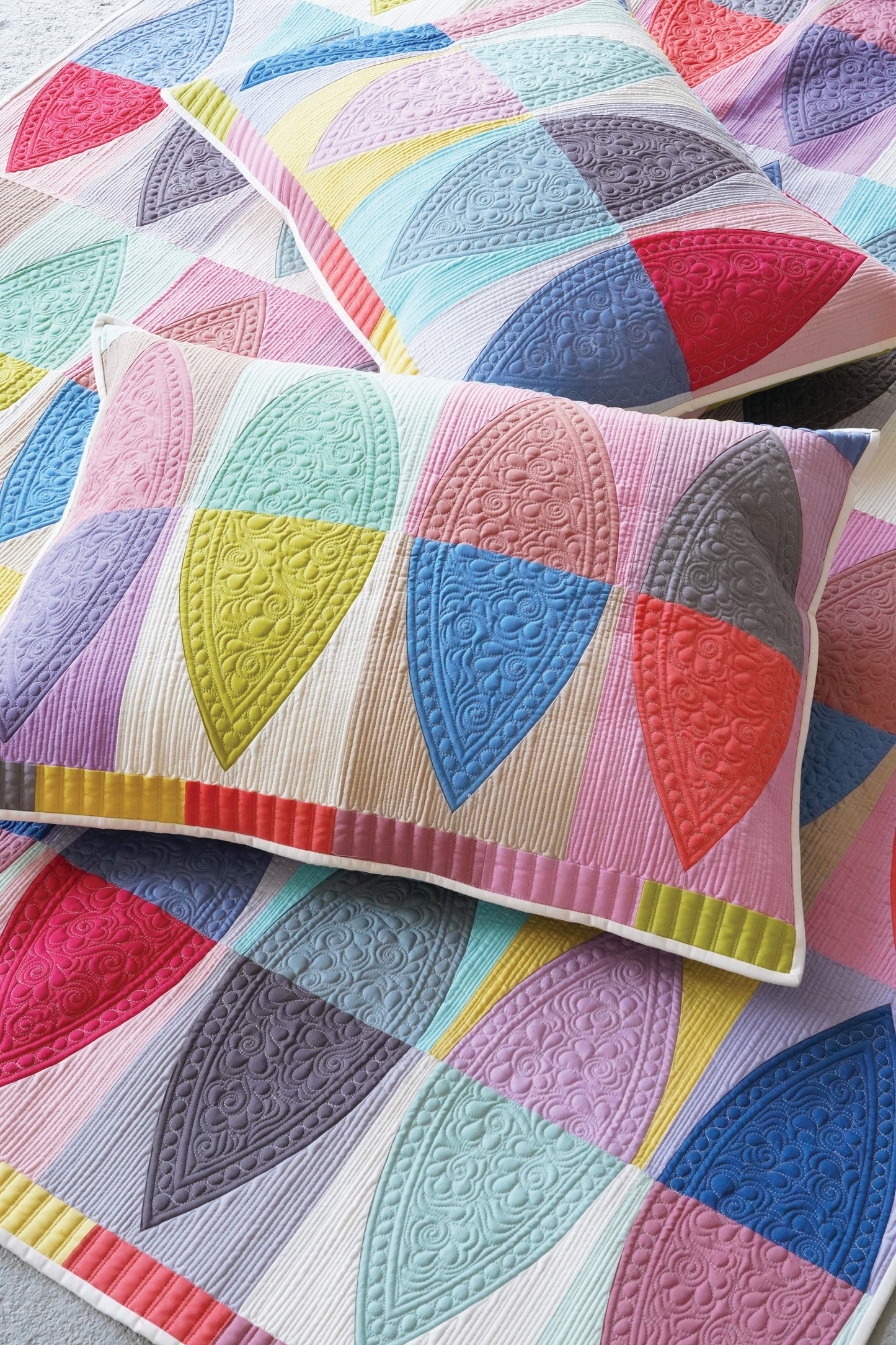 A close-up of a quilted throw pillow and matching quilt featuring a colorful geometric patchwork design with leaf-like shapes in pastel pink, blue, yellow, purple, and coral on a white base.
Photo by Tildafabrics.com [additional_image_link]