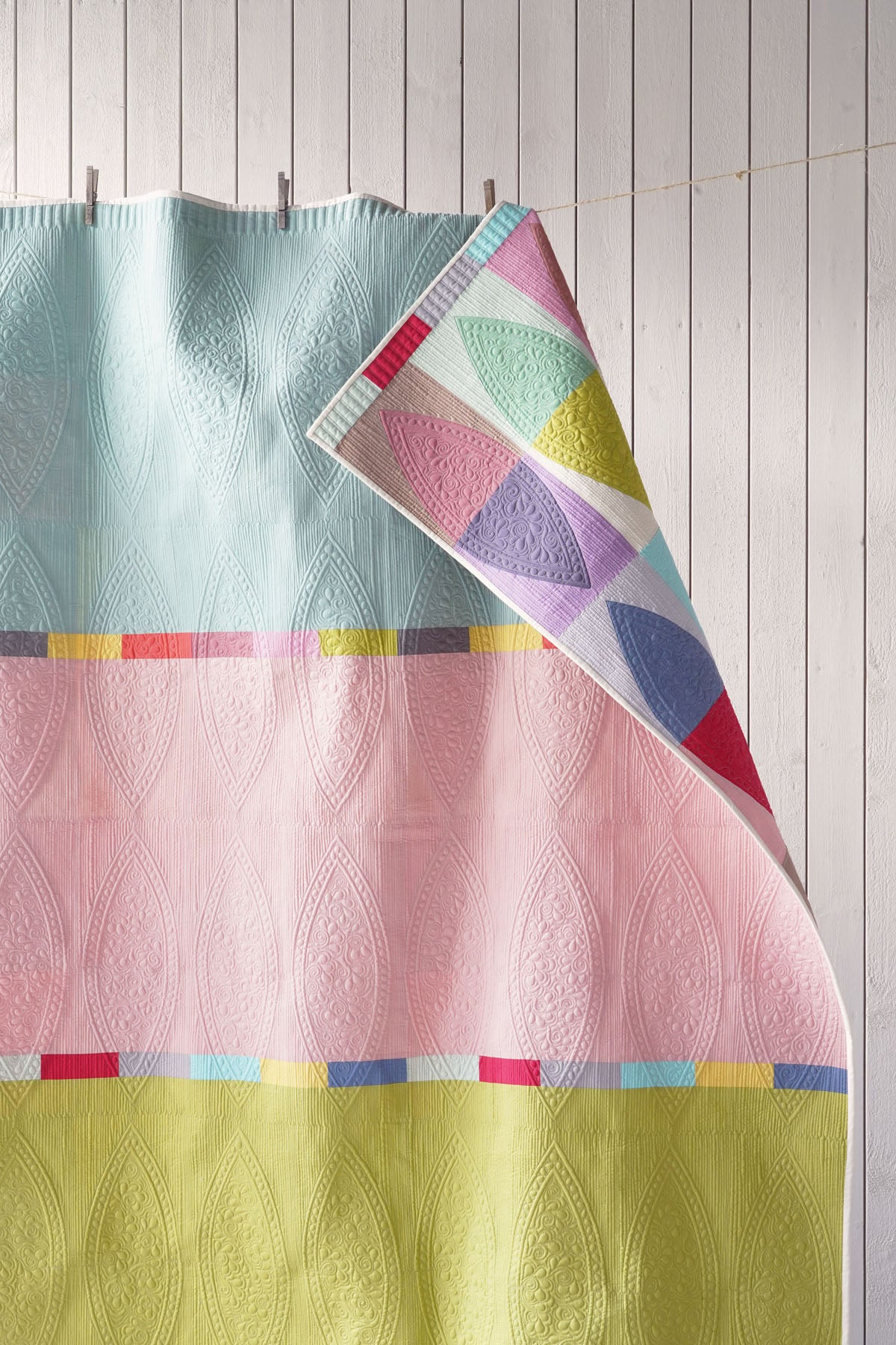 A pastel quilt hanging against a white paneled wall, with one corner folded back to show a geometric multicolor pattern. The main visible color areas are pale aqua, pink, and pale yellow with thin multicolor stripe accents.
Photo by Tildafabrics.com [additional_image_link]