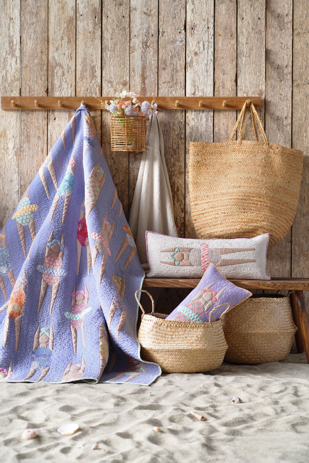 A styled beach-themed home decor display featuring a lavender quilted blanket with a colorful ice cream cone pattern draped over a rustic wooden bench, alongside woven straw baskets, a tote bag, and matching pillows on a sandy surface against a wood plank wall.
Photo by Tildafabrics.com [additional_image_link]