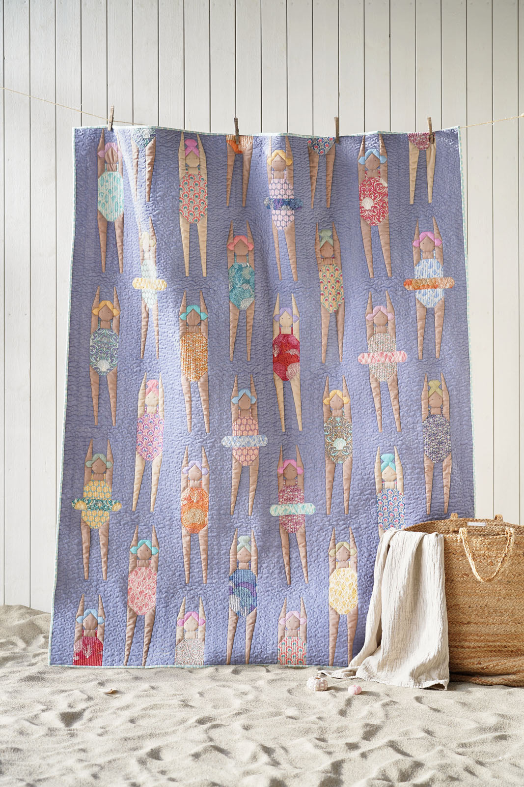 A beach-themed quilt or blanket featuring a repeating pattern of women in swimsuits on a lavender-blue background, styled on a sandy floor against a white wood-paneled wall with a straw bag and white towel nearby.
Photo by Tildafabrics.com [additional_image_link]