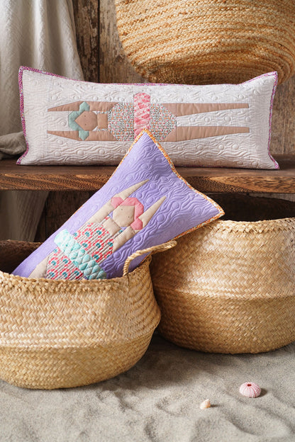 Two decorative throw pillows displayed in woven baskets on a rustic wooden bench. The back pillow is white with a pink mermaid design, and the front pillow is lavender with a multicolor swimmer design.
Photo by Tildafabrics.com [additional_image_link]