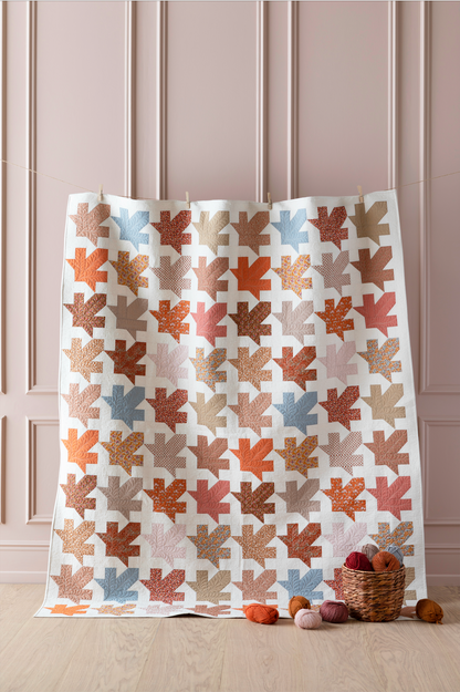 A quilt with a maple leaf pattern featuring various shades of autumn such as brown, orange, and yellow, displayed on a hanger.
Photo by Tildafabrics.com [image_link]