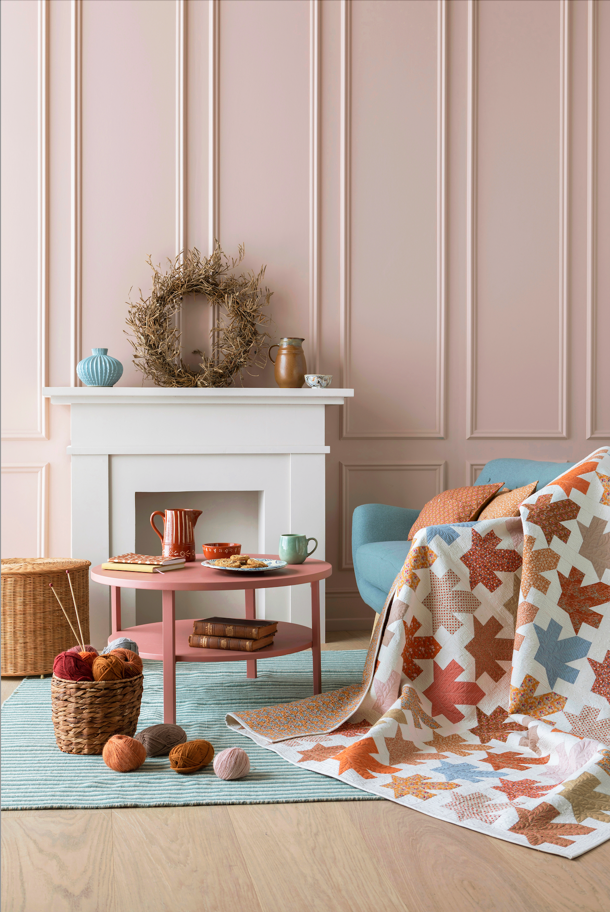 A cozy living room scene with a patterned throw blanket draped over a light blue armchair beside a pink coffee table and a white fireplace mantel. The blanket has a white base with a geometric houndstooth-like pattern in terracotta, rust, tan, and muted blue.
Photo by Tildafabrics.com [additional_image_link]