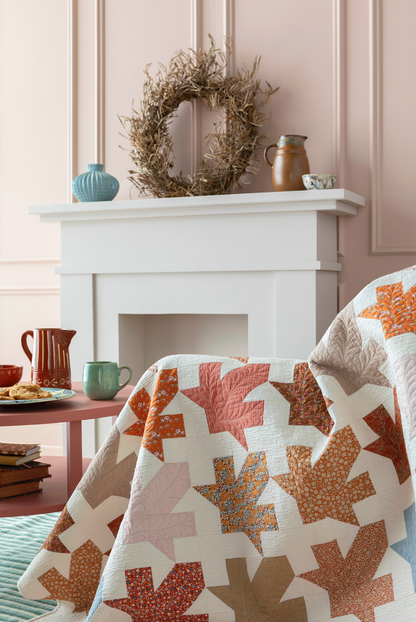 A quilted throw blanket with a geometric patchwork pattern of orange, rust, beige, and white leaf-like shapes is draped over the front of a sofa in a styled living room setting with a white fireplace and decorative wreath.
Photo by Tildafabrics.com [additional_image_link]
