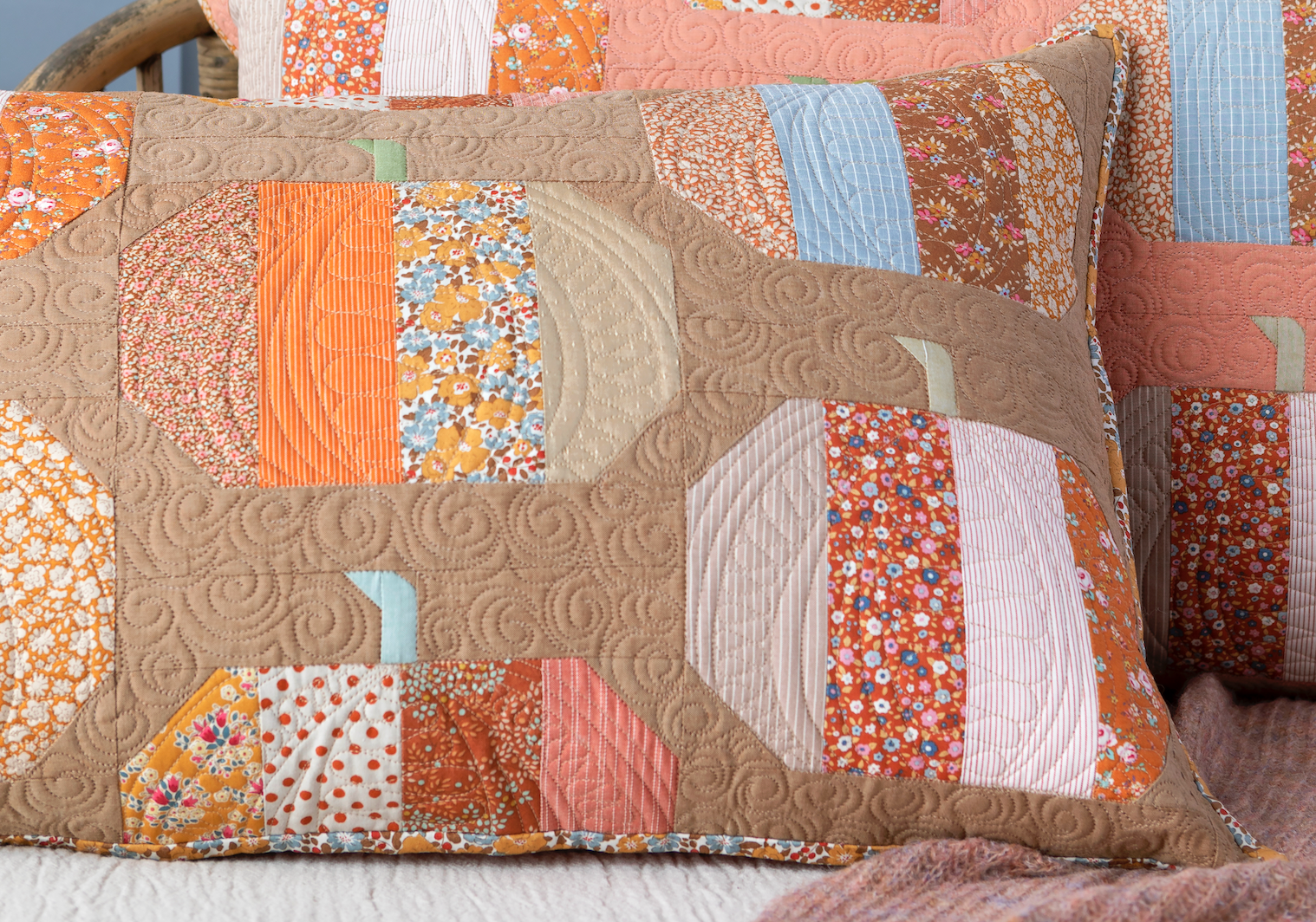A close-up of a quilted patchwork pillow featuring autumn-themed fabric panels in orange, beige, pink, and light blue, with a matching quilted pillow behind it.
Photo by Tildafabrics.com [image_link]