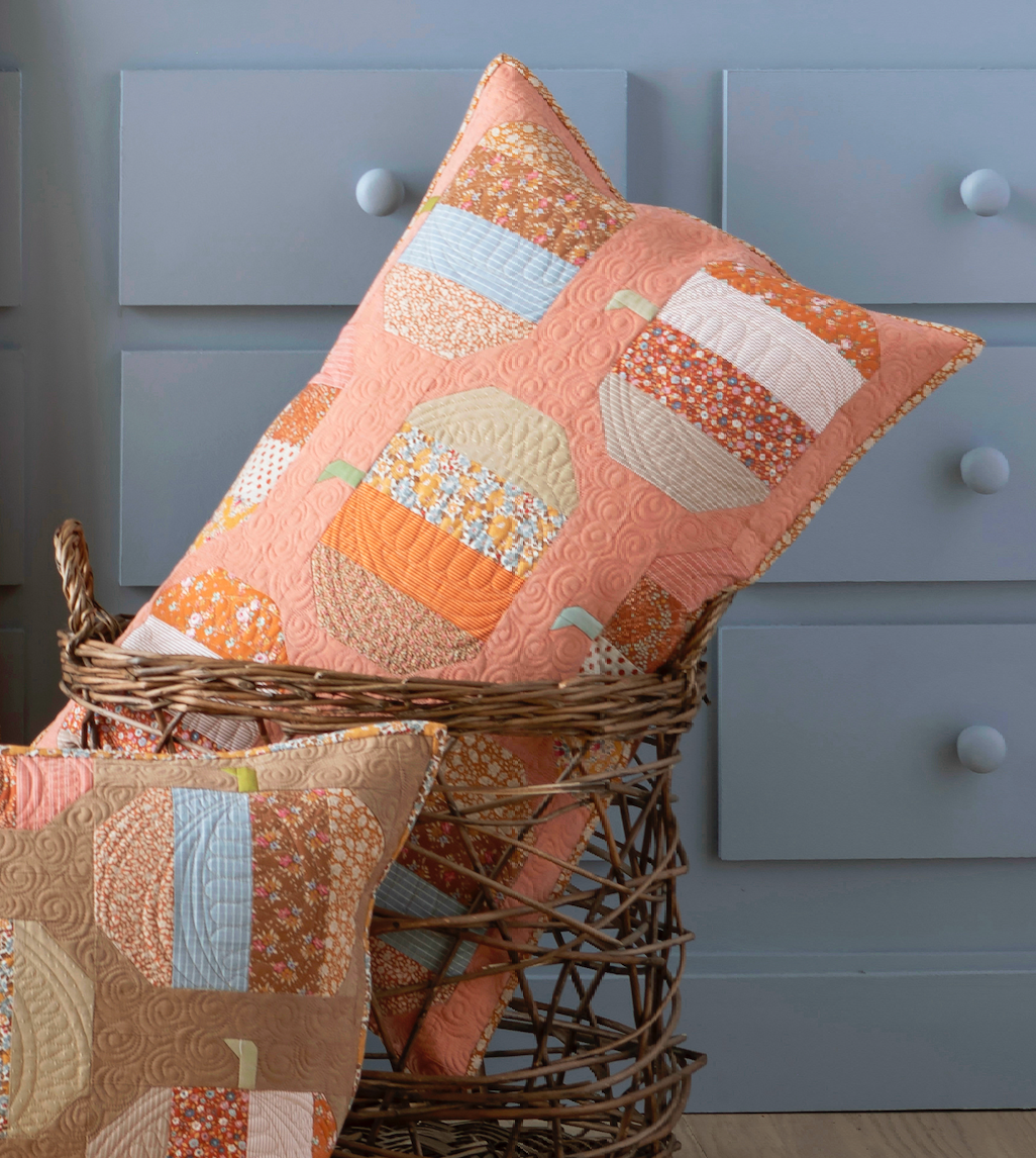 Two quilted throw pillows with a patchwork pumpkin design in peach, orange, beige, and light blue, styled in a wicker basket in front of a blue-gray dresser.
Photo by Tildafabrics.com [additional_image_link]