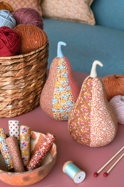 Two decorative pear-shaped fabric ornaments with floral patterns and stitched stems, displayed on a pink table alongside yarn, thread rolls, knitting needles, and a wicker basket in a cozy craft setting.
Photo by Tildafabrics.com [additional_image_link]