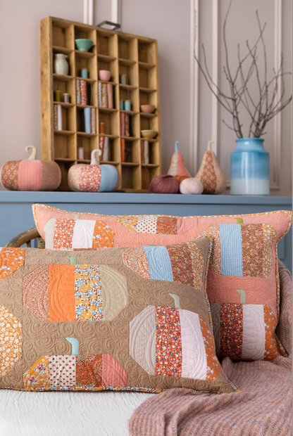 Three decorative throw pillows with a patchwork pumpkin design in warm autumn colors, styled on a bed with a pink blanket, with a wooden shelf and blue cabinet in the background.
Photo by Tildafabrics.com [additional_image_link]