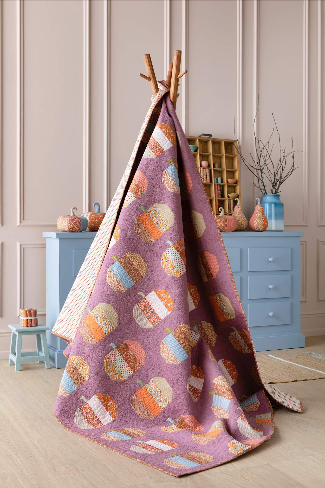 A fabric teepee play tent with a purple base, decorated with a pattern of colorful pumpkins, displayed on a wooden stand in a child's playroom setting.
Photo by Tildafabrics.com [additional_image_link]