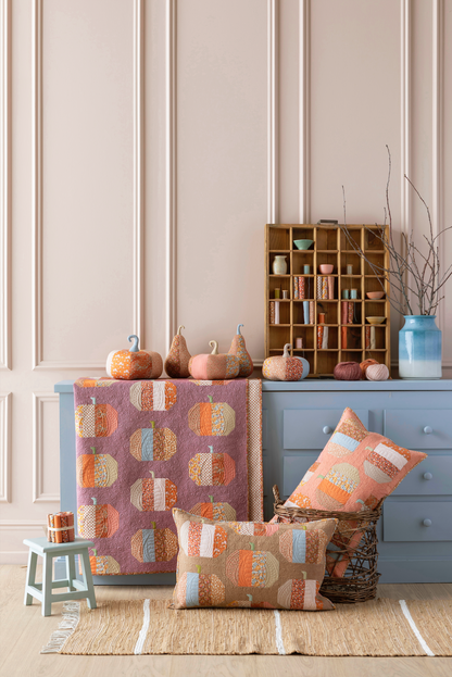 A styled home decor scene showing a quilted throw blanket and matching pillows with a pumpkin patch pattern in orange, beige, and light blue, displayed on a blue dresser in a room with paneled walls and autumn decor.
Photo by Tildafabrics.com [additional_image_link]
