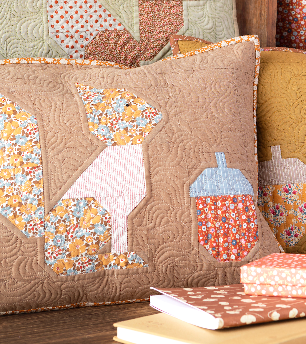 A quilted throw pillow in a warm tan color with a patchwork design of acorns and leaves in floral and striped fabrics, styled on a wooden surface with other quilts and books in the background.
Photo by Tildafabrics.com [image_link]